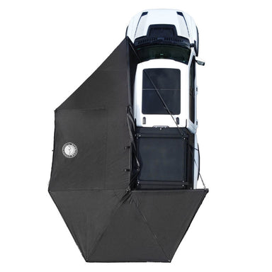 Top-down view of a white SUV with an OVS Nomadic Awning partially deployed. The black awning is attached to the side of the vehicle, extending outwards and downwards to create a covered space