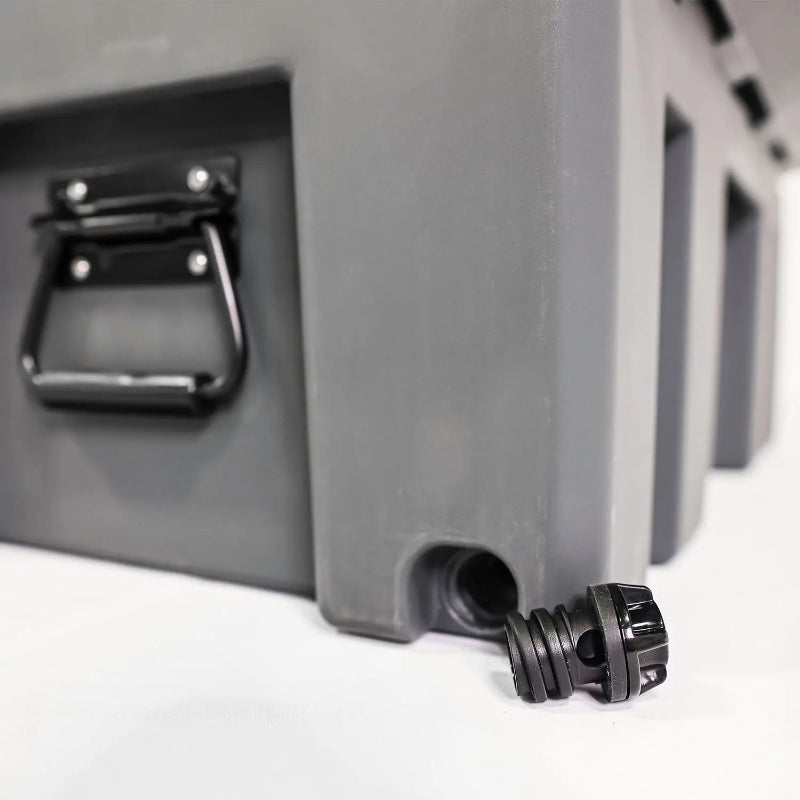 Image of a close-up of a black drain plug next to a grey storage box with a side handle, angled side view on a white background.