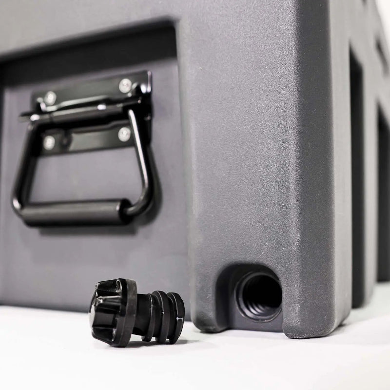 Image of a close-up of a black drain plug next to a grey storage box with a side handle, angled side view on a white background.