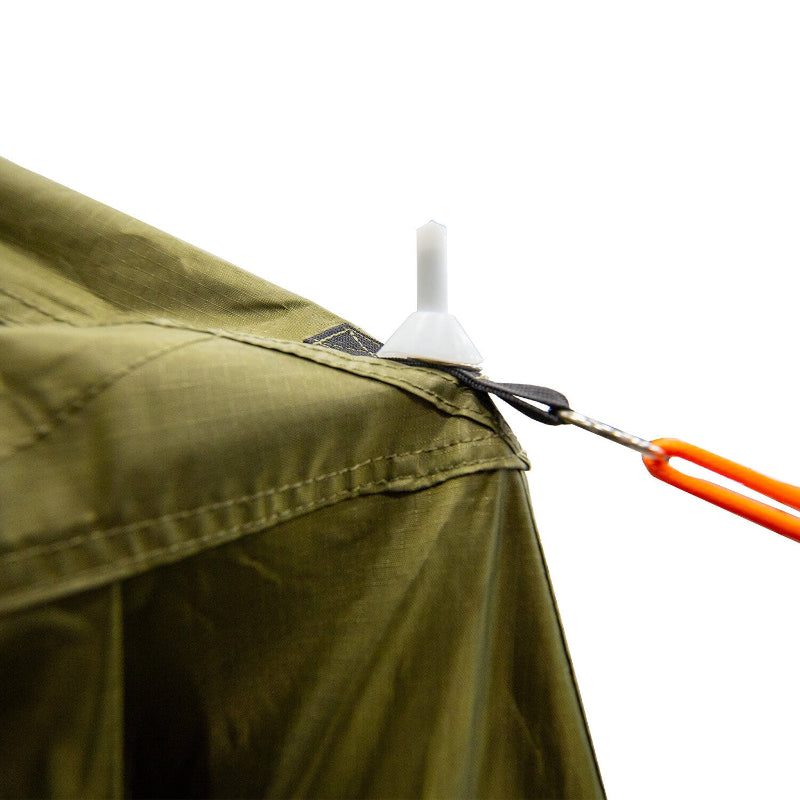 Image of a close-up detail of the 23ZERO Annex Armadillo A3 Plus tent, showing a white stake or anchor point attached to the olive-green tent fabric with an orange pull strap, angled view on a white background