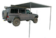 Image of a black Peregrine Awning partially deployed, extending from the side of a dark grey Nissan SUV. The awning is supported by two black poles, providing shade, and is shown against a white background