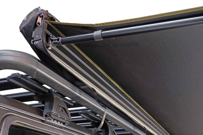 Close-up image of a Peregrine Awning partially deployed, showcasing the black underside of the fabric and the structural poles. The awning is attached to a vehicle's roof rack, with the rolled-up cover visible at the far end, all against a white background