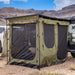 Image of a green Bushman awning room fully set up with black mesh windows, attached to an off-road trailer in a rugged, mountainous desert landscape. A person is visible inside the awning room, and camping gear is nearby