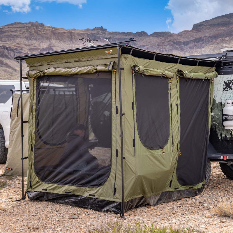 Image of a green Bushman awning room fully set up with black mesh windows, attached to an off-road trailer in a rugged, mountainous desert landscape. A person is visible inside the awning room, and camping gear is nearby