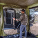 Image of a man in a plaid shirt and cap opening the door of a black off-road trailer from inside a green Bushman awning room. The interior of the awning room and a portion of the trailer with "PEDALTON TRAILERS" branding are visible