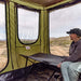 Image of a woman seated on a cot inside a green Bushman awning room, looking out at a desert landscape with mountains under a partly cloudy sky. The awning room's mesh window is open, offering an expansive view.
