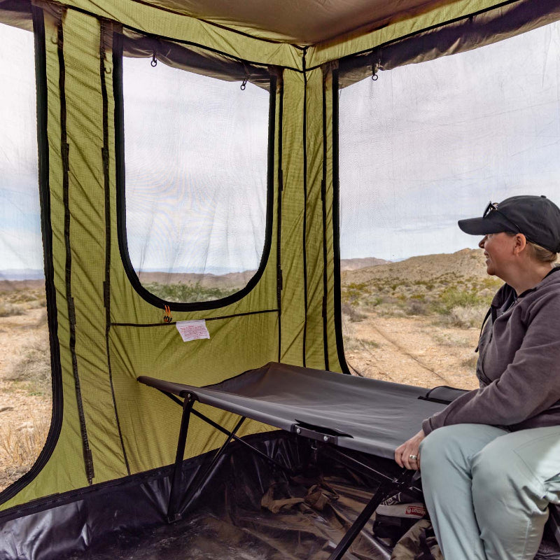 Image of a woman seated on a cot inside a green Bushman awning room, looking out at a desert landscape with mountains under a partly cloudy sky. The awning room's mesh window is open, offering an expansive view.
