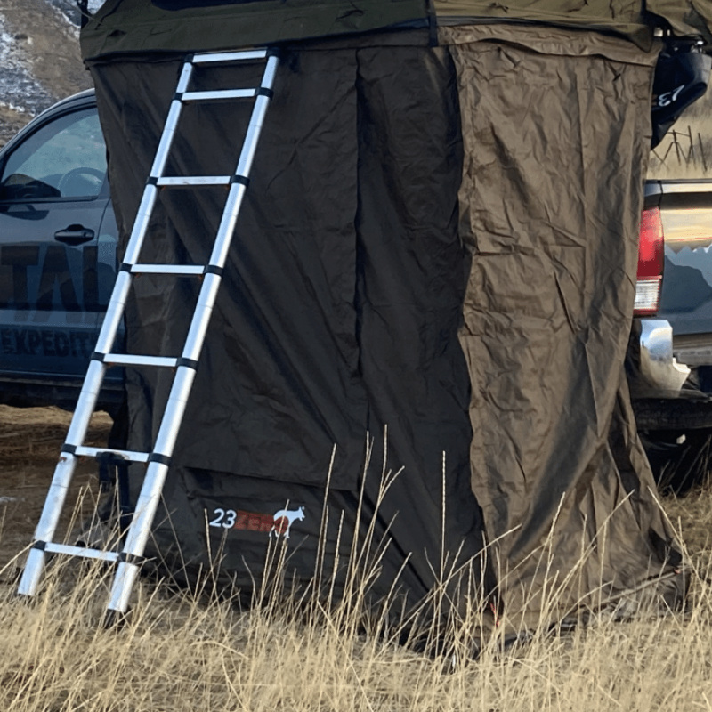 Image of a brown rooftop tent with an attached black awning room and a silver ladder, set up next to a truck in a dry, grassy field. The "23 ZERO" logo is visible on the awning room