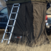Image of a brown rooftop tent with an attached black awning room and a silver ladder, set up next to a truck in a dry, grassy field. The "23ZERO" logo is visible on the awning room