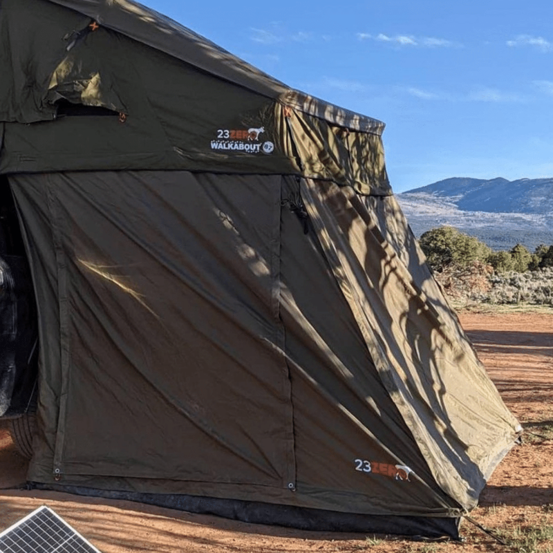 Image of a green Walkabout Rooftop Tent Annex set up on a dirt campsite with mountains in the background. The tent annex is fully deployed, and a portable solar panel can be seen in the bottom left corner of the image