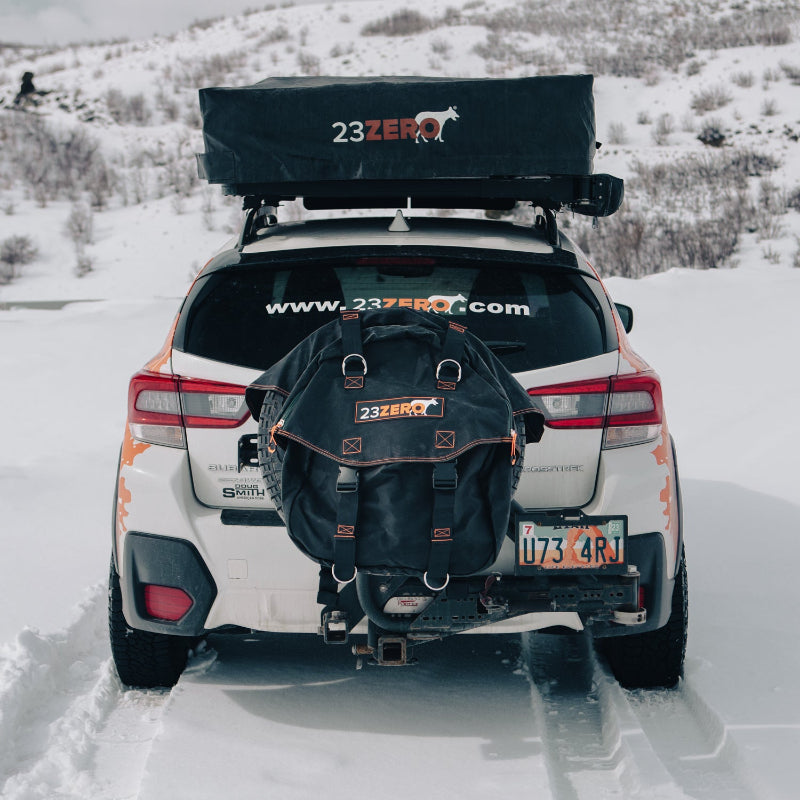 Image of the rear of a white Subaru Crosstrek with a black 23ZERO rooftop tent and a black 23ZERO spare tire storage bag, parked in a snowy landscape.