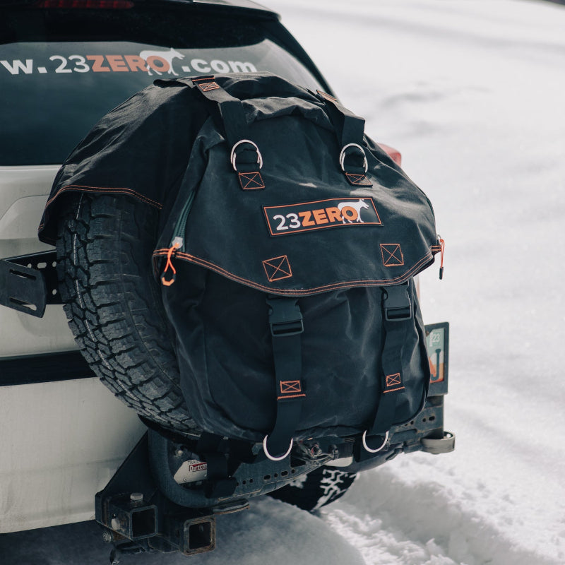 Image of a black 23ZERO spare tire storage bag with orange stitching and straps, mounted on the rear spare tire of a white vehicle, set against a snowy outdoor background.
