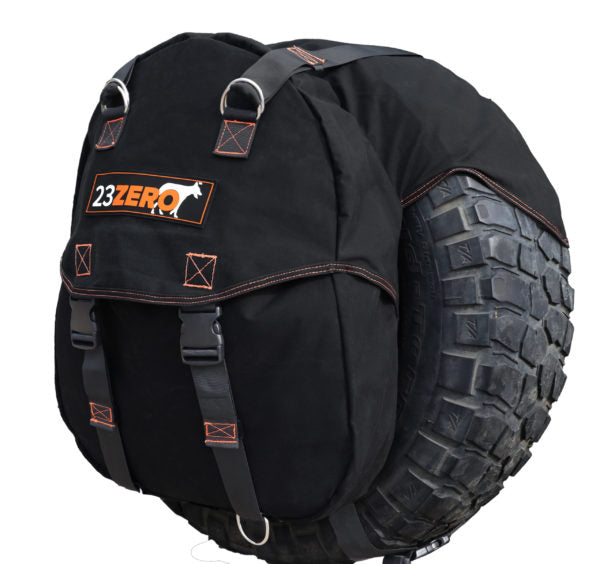 Image of a black 23ZERO spare tire storage bag with orange stitching, black straps, and metal hardware, mounted on a rugged tire, viewed at a slight angle on a white background.