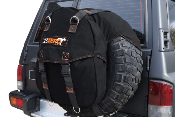 Image of a black 23ZERO spare tire storage bag with orange stitching and black straps, mounted on the rear spare tire of a dark grey SUV, shown from a slight angle.