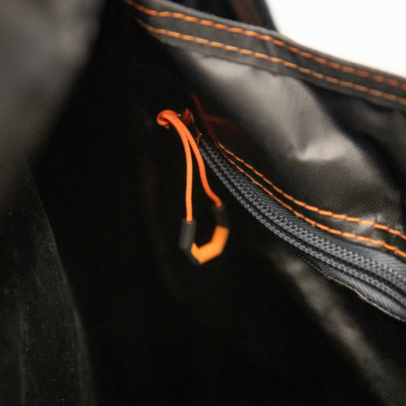 Image of the interior of a black bag, showing an open zipper with an orange pull tab and orange stitching along the fabric edge.