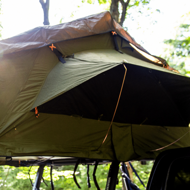 Image of a green roof top tent with an awning partially deployed, with a dark underside and bright highlights, angled from the front and side, set against a blurred green forest background.