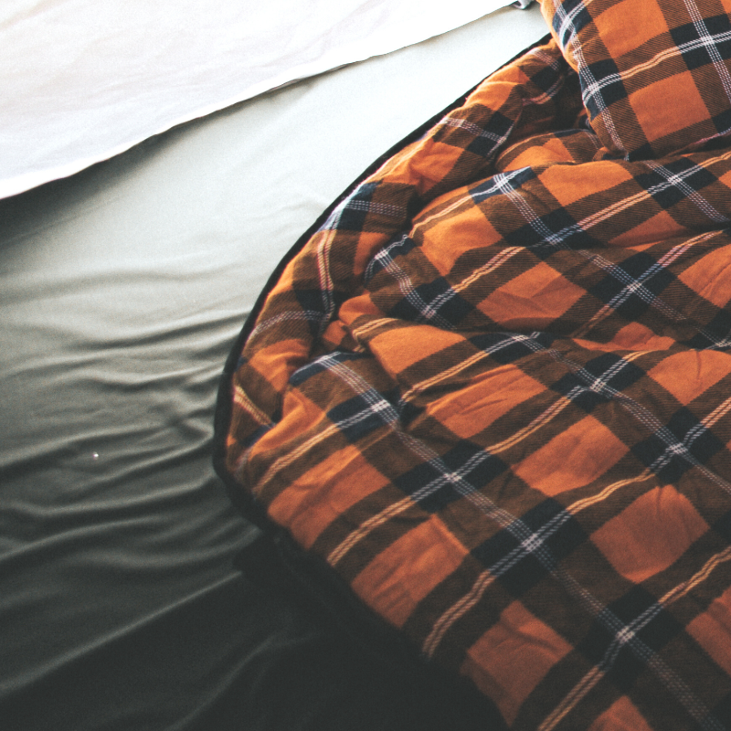 Image of Kabari sleeping bag partially spread on a bed, orange and black plaid insulated fabric with visible stitching, angled top and side view on a light grey sheet background.