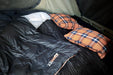 Image of a 23ZERO Camping Quilted Blanket in dark grey with orange trim, laid out with a white pillow and two orange plaid pillows inside a tent, ready for use.