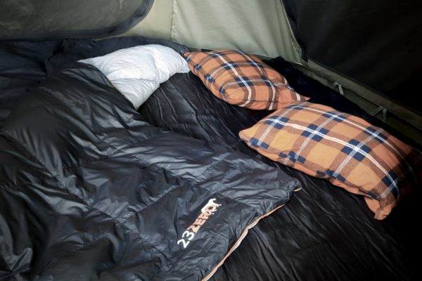 Image of a 23ZERO Camping Quilted Blanket in dark grey with orange trim, laid out with a white pillow and two orange plaid pillows inside a tent, ready for use.