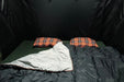 Image of a 23ZERO Camping Quilted Blanket, dark grey on one side and light grey with orange trim on the other, laid out on a green sleeping pad with two orange plaid pillows inside a dark tent.