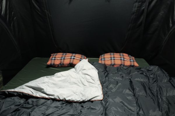 Image of a 23ZERO Camping Quilted Blanket, dark grey on one side and light grey with orange trim on the other, laid out on a green sleeping pad with two orange plaid pillows inside a dark tent.