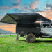 Image of a gray pickup truck with a black bed canopy and a black 23ZERO Peregrine Pro 270-degree awning fully deployed, parked in a grassy field beside a lake with mountains in the background under a sunset sky. A bicycle is leaning against the truck's canopy
