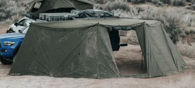 Image of a Peregrine 180-degree Awning with two walls attached, set up next to a white pickup truck in a dry, grassy landscape. The green canvas awning and walls are fully extended to create an enclosed living space, with an open doorway on the right.