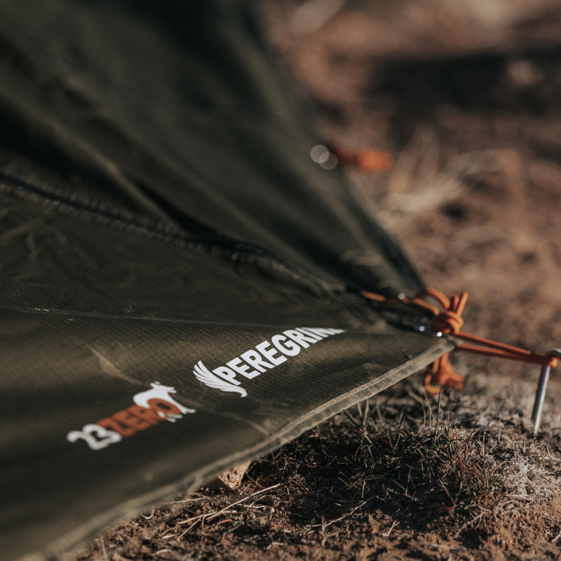 Close-up image of the Peregrine 270 Awning's green fabric touching the ground, with the "23ZERO PEREGRINE" logo prominently displayed in white. An orange tie-down rope and stake are visible in the foreground, securing the awning to the earthy terrain