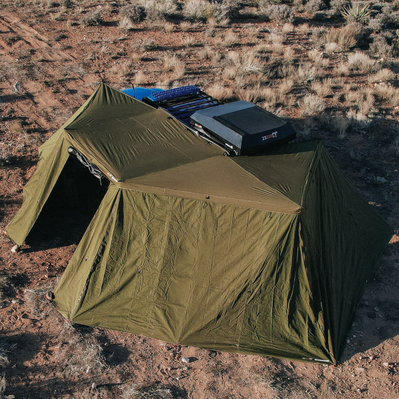Aerial image of a blue pickup truck with a rooftop tent and a fully deployed green Peregrine 270 Awning with all walls attached, forming an enclosed room. The setup is situated on dry, brush-covered ground under natural light