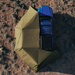 Aerial image of a blue pickup truck with a roof rack and a rooftop tent, connected to a fully deployed green Peregrine 270 Awning. The awning extends outward from the truck, providing extensive shaded coverage over rough, reddish-brown terrain