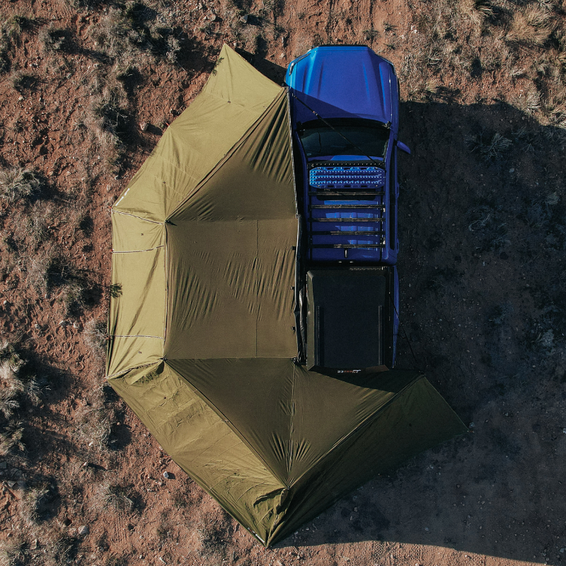 Aerial image of a blue pickup truck with a roof rack and a rooftop tent, connected to a fully deployed green Peregrine 270 Awning. The awning extends outward from the truck, providing extensive shaded coverage over rough, reddish-brown terrain
