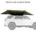 Image of a 23ZERO Peregrine 180 Compact Awning partially deployed from the rear of a vehicle. The awning features green fabric with a black underside and is mounted on a white vehicle with a roof rack