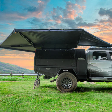 Image of 23Zero Peregrine Pro 270° 5-arm awning partially deployed on a black vehicle, with a scenic sunset background, mounted on a rugged off-road truck with bicycle attached