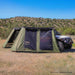 Image of a green 23ZERO Peregrine wall kit attached to an awning, creating an enclosed room next to a white Jeep Rubicon. The wall kit features large mesh screen windows and is set up in a sunny, dry, grassy landscape with rolling hills in the background