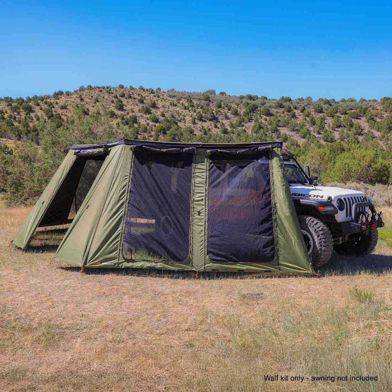 Image of a green 23ZERO Peregrine wall kit attached to an awning, creating an enclosed room next to a white Jeep Rubicon. The wall kit features large mesh screen windows and is set up in a sunny, dry, grassy landscape with rolling hills in the background