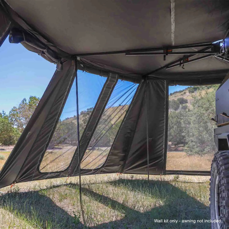 Image of the interior of a 23ZERO Peregrine wall kit, viewed from inside the enclosed space. The dark green walls have large, clear plastic windows, and the black awning structure is visible overhead, with a dirt road and hills in the background