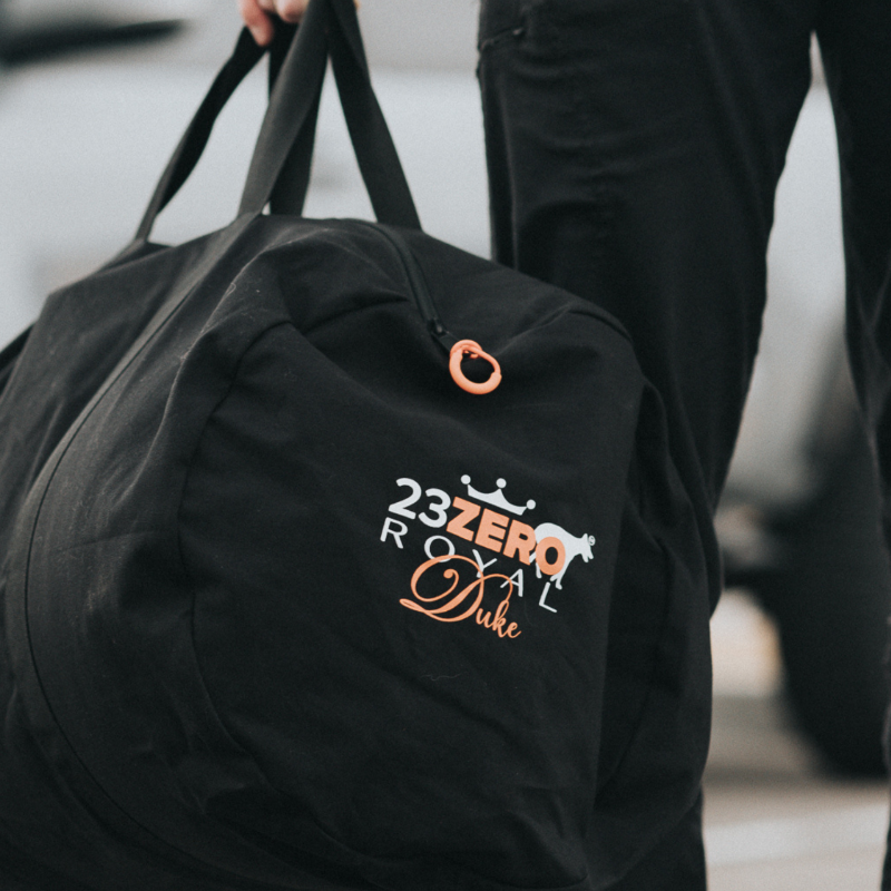 Image of a person holding a black 23ZERO Royal Duke branded duffel bag with an orange zipper pull tab, with part of their leg visible in black pants.