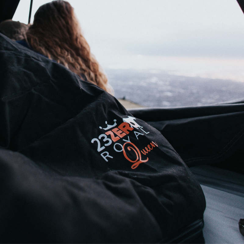 Image of a black sleeping bag with a "23ZERO Royal Queen" logo, seen from inside a tent, with a person's hair and an outdoor view in the blurred background.