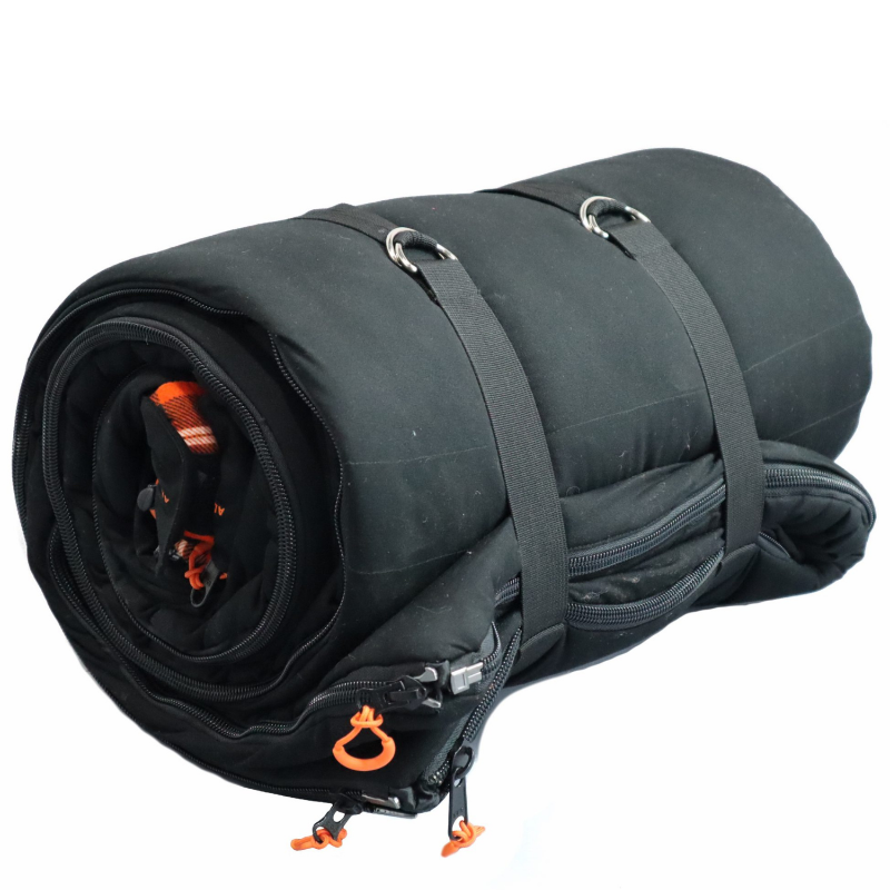 Image of a black sleeping bag, tightly rolled and secured with black straps and metal D-rings, showcasing its zippers with orange pull tabs and a glimpse of an orange plaid interior, all against a white background.