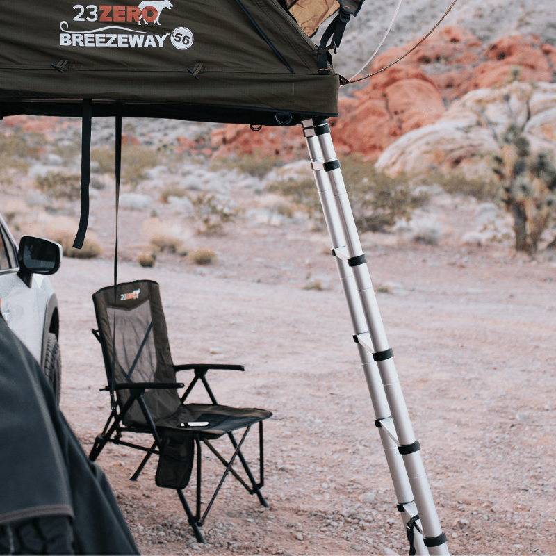 Image of a dark green roof top tent deployed from a vehicle, with a telescopic ladder extended to the ground and a black camping chair positioned underneath, all set in a desert landscape.