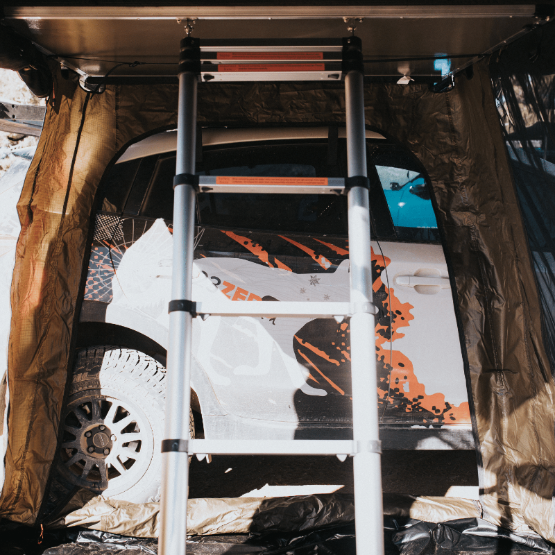 Image showing a silver telescopic ladder leading up to a roof top tent, with a white vehicle featuring orange graphics visible through the tent's annex, set against a bright, sunny background.