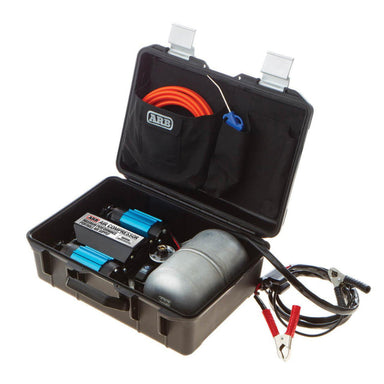 Image of an ARB Twin Motor Portable 12V Air Compressor (CKMTP12) kit, opened to display its contents: a black hard case containing two blue air compressors, a silver air tank, an orange air hose, and red and black battery clamps, all ready for use
