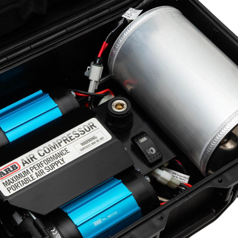 Image of the interior of an ARB portable air compressor kit, showing a close-up of the black compressor unit with blue cooling fins and a "MAXIMUM PERFORMANCE PORTABLE AIR SUPPLY" label, connected to a cylindrical silver air tank, all housed within a black hard case. Wires and a pressure gauge are also visible
