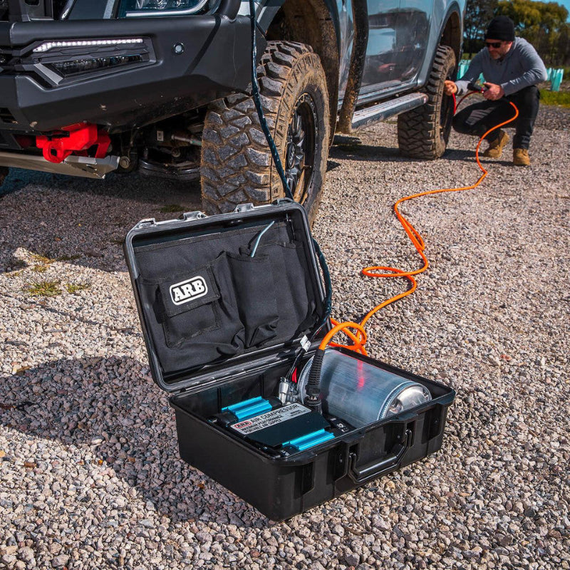 Image of an ARB portable air compressor kit, open and placed on a gravel ground, connected via an orange hose to the tire of a gray off-road truck. A man is kneeling near the truck's front tire, operating the air hose