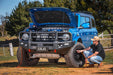 Image of a man airing up a tire on a blue Ford Bronco with the hood open, showcasing aftermarket modifications in a rural outdoor setting