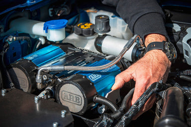 Image of a person's hand adjusting a blue ARB Brushless air compressor mounted in a vehicle's engine bay, showing various hoses and fluid reservoirs