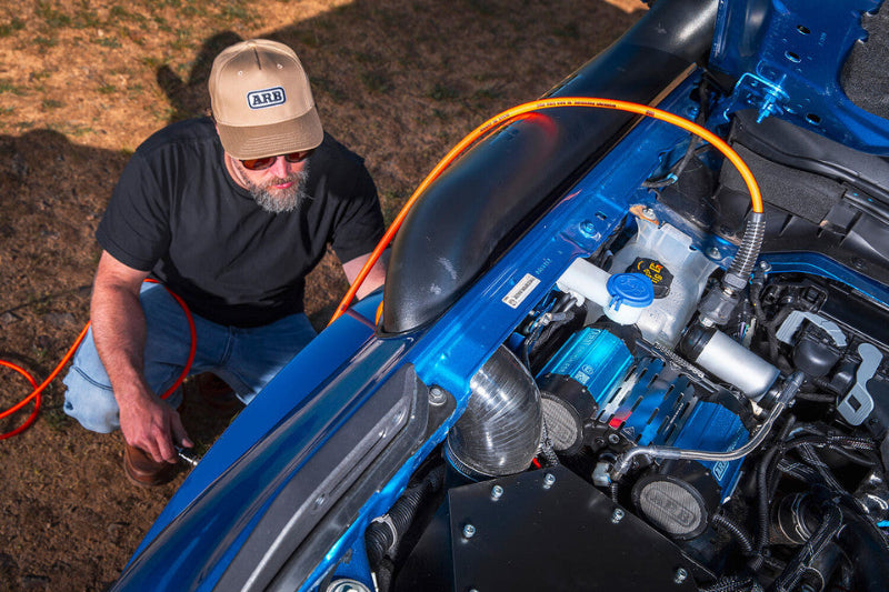 Image of a man in an ARB cap and sunglasses operating an air hose from a blue ARB air compressor mounted in the engine bay of a blue vehicle, in an outdoor setting
