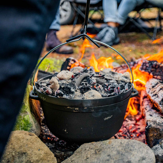 Image of cast iron camp Dutch oven hanging over open fire with hot coals on lid, outdoor camping scene with flames and rocks in background.