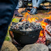 Image of cast iron camp Dutch oven hanging over open fire with hot coals on lid, outdoor camping scene with flames and rocks in background.