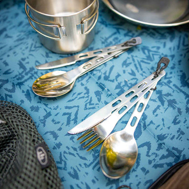 Image of GSI stainless steel 3-piece camping cutlery sets with spoon, fork, and serrated knife laid out on blue patterned camp table beside metal cookware, angled top view.